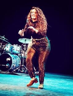 Tap artist Michela Lerman dances on a turquoise-lit stage, with a drumset and a tenor saxophonist in the background.